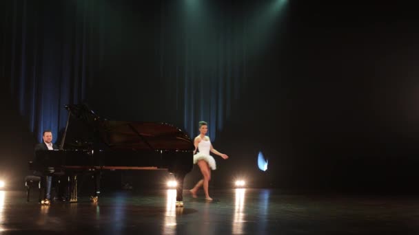 männlicher Pianist am Flügel und tanzende Ballerina im weißen Kleid auf dunkler Bühne