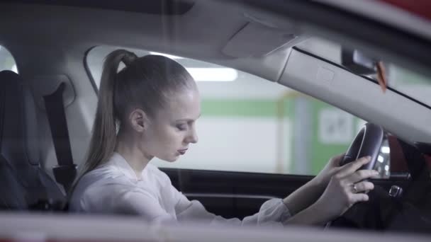 Pretty woman in white shirt sits in car, holds steering wheel next to garage — Stock Video