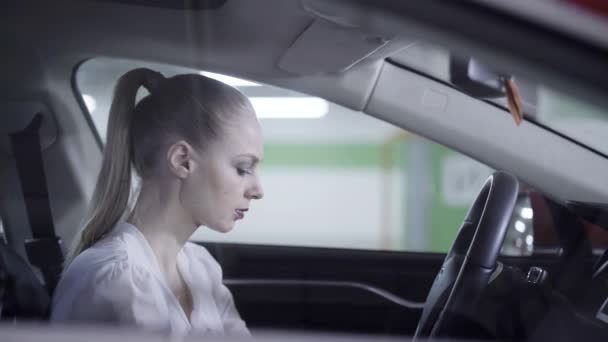 Young female in white shirt sits in car, holds steering wheel next to garage. — Stock Video