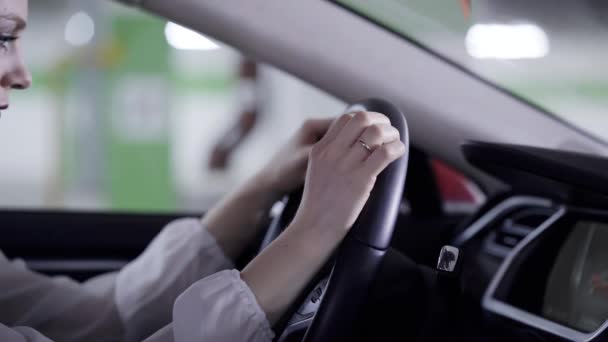 Mooi meisje in wit overhemd zit in de auto, houdt van stuurwiel naast garage — Stockvideo