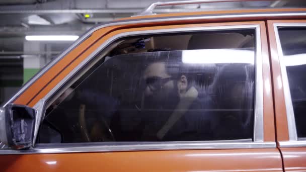 Молодий бородатий хлопець з довгим темним волоссям відкриває вікно червоного автомобіля і дивиться з нього — стокове відео
