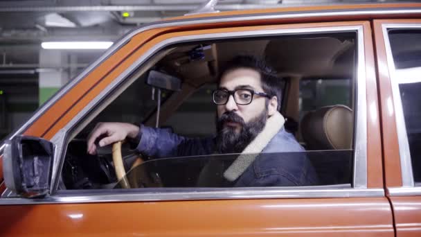 Симпатичный бородатый парень с длинными темными волосами открывает окно красной машины и смотрит из нее — стоковое видео