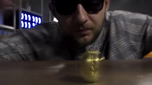 サングラスでかわいい若い男性は茶色のテーブルの上に大きな新しい金ビットコインコインをスピン. — ストック動画