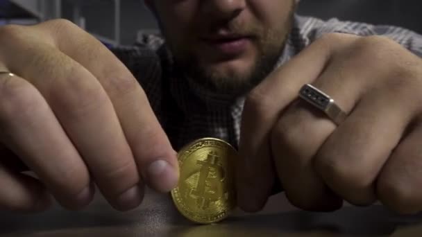Young man in sunglasses joyfully spins big new gold bitcoin coin on brown table. — Stock Video