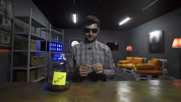 Smiling guy blogger drops big gold bitcoin into glass jar in front of camera — Stock Video