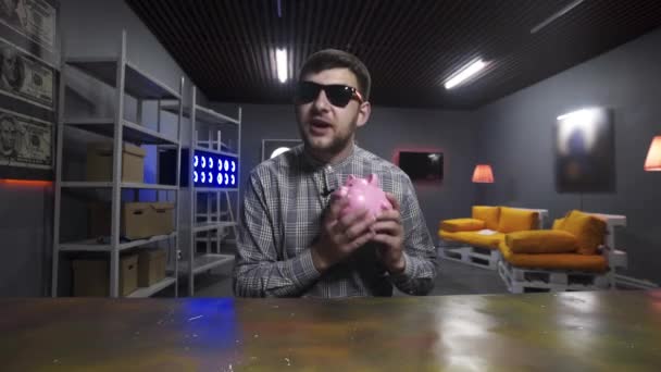 Bearded man sits behind long table with piggy bank in hand and speaks at camera — Stock Video