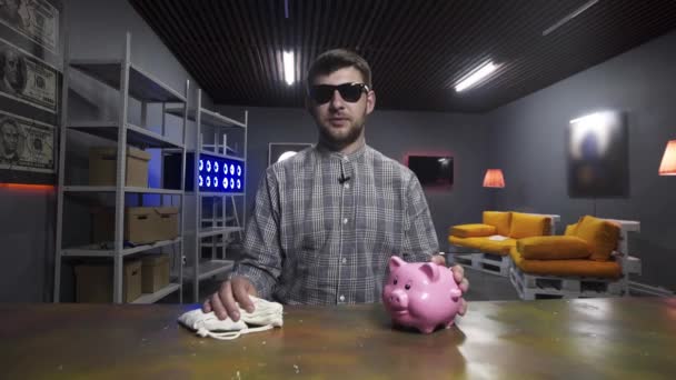 Young handsome man speaks and looks at camera holding piggy bank in hands — 비디오