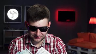 Güneş gözlüklü beyaz adam konuşuyor, kameraya bakıyor, cüzdanını parasıyla atıyor.