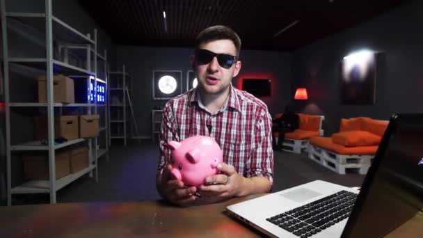 Привлекательный мужчина в солнечных очках держит пластиковую свинью, говорит и смотрит в камеру — стоковое видео