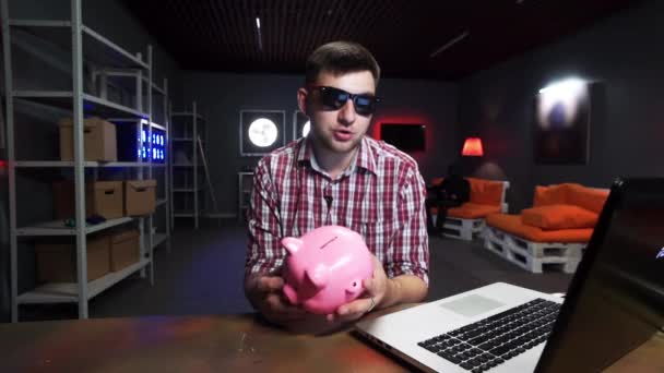 Joven hombre activo con gafas de sol sostiene cerdo plástico, habla y mira a la cámara — Vídeos de Stock