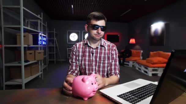 Young bearded male sits behind table with laptop, holds plastic pig and speaks — Stock Video