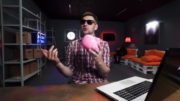 Young bearded man with sunglasses holds plastic pig, speaks and looks at camera — Stockvideo