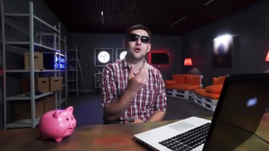 Güneş gözlüğü ile Çekici genç adam masada domuz ve dizüstü bilgisayar olan kamera konuşuyor