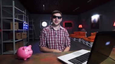 Güneş gözlüğü ile Sakallı genç adam masada domuz ve dizüstü bilgisayar olan kamera konuşuyor