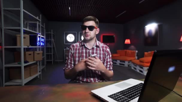 Lustiger junger Mann mit Sonnenbrille reibt sein Handy mit Hemd ein und spricht in die Kamera — Stockvideo