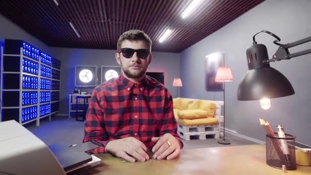 Homem bonito barbudo senta-se atrás da mesa, mostra a língua no quarto com refrigeradores iluminados — Vídeo de Stock