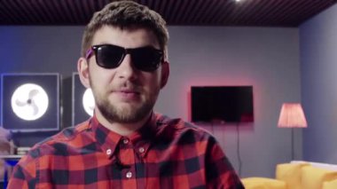 Komik adam konuşuyor ve mobilyalı odada kameraya bakarak el hareketleri kullanır.