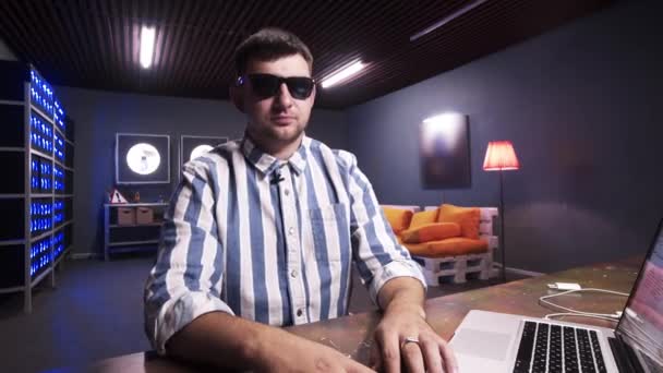 Glimlachend volwassen man is vloggen zittend achter tafel met laptop in donkere kamer — Stockvideo