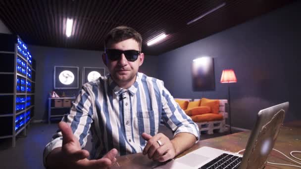 Beau gars parle à la caméra assis derrière le bureau avec ordinateur portable dans la chambre grise — Video
