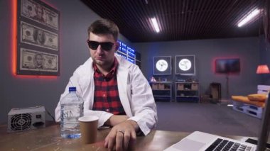 Beyaz ceketli güneş gözlüklü adam plastik şişe ve bardak ile masanın arkasında oturur