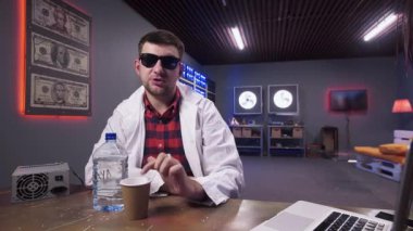 Beyaz ceketli sevimli sakallı adam plastik şişe ve fincan ile masanın arkasında oturur
