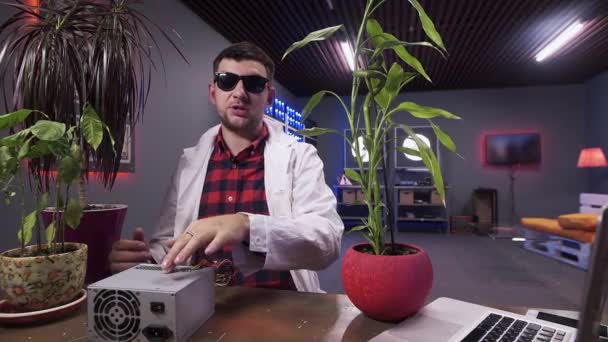 Facet w białym płaszczu laboratoryjnym przemawia do kamery za biurkiem z niektórymi roślinami w misach. — Wideo stockowe