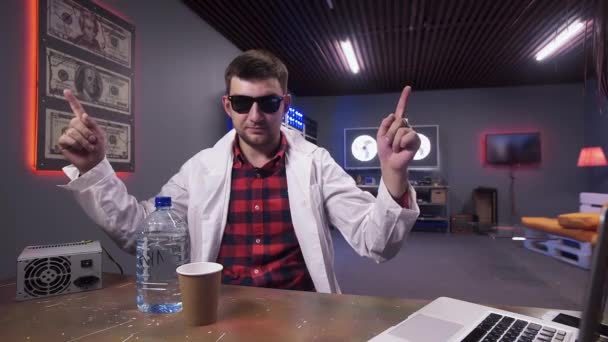 Guapo joven con bata de laboratorio blanca se sienta detrás del escritorio con botella de plástico — Vídeos de Stock