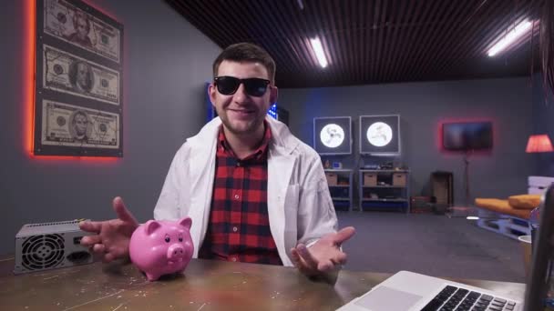 Человек ученый в солнцезащитных очках и халате говорит, имея свинью и ноутбук на столе — стоковое видео