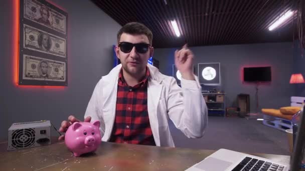 Hombre con gafas de sol y bata de laboratorio blanca habla, teniendo cerdo y portátil en el escritorio — Vídeos de Stock