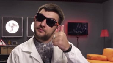 Güneş gözlüğü ve beyaz laboratuvar önlüğü giyen yakışıklı sakallı adam başparmak kadar gösterir.