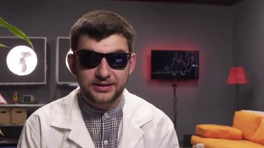 Güneş gözlüğü ve beyaz laboratuvar önlüğü giyen sakallı erkek bitki ile pot tutarak konuşuyor