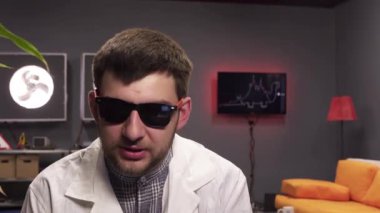 Güneş gözlüğü ve beyaz laboratuvar önlüğü giyen güzel adam bitki ile pot tutarak konuşuyor.