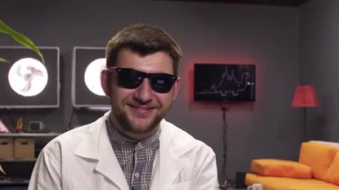 Güneş gözlüğü ve beyaz laboratuvar önlüğü giyen sevimli genç erkek sevinçle gülüyor.