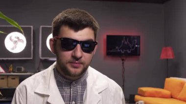 Sakallı, güneş gözlüğü ve beyaz laboratuvar önlüğü giyen çekici adam sevinçle konuşuyor.