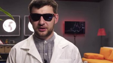 Güneş gözlüğü ve beyaz laboratuvar önlüğü giyen iyi görünümlü genç adam sevinçle konuşuyor.