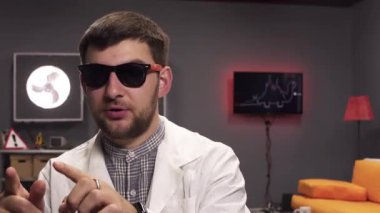 Güneş gözlüğü ve beyaz laboratuvar önlüğü giyen sakallı adamın yüzü sevinçle konuşuyor.