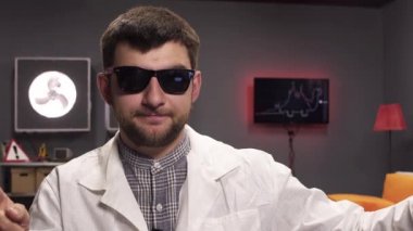 Güneş gözlüğü ve beyaz laboratuvar önlüğü giyen sakallı adamın yüzü sevinçle konuşuyor.