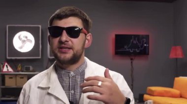 Güneş gözlüğü ve beyaz laboratuvar önlüğü giyen sakallı sevimli adam sevinçle konuşuyor.