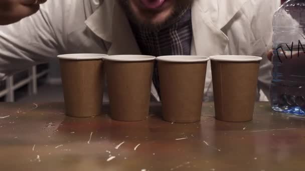 白いラボコートを着たひげを生やした男が4つの紙コップから水を舐めようとする. — ストック動画