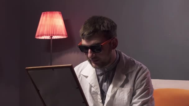 Hombre caucásico con barba usando gafas de sol y laboratorio blanco sostiene imagen en marco — Vídeo de stock