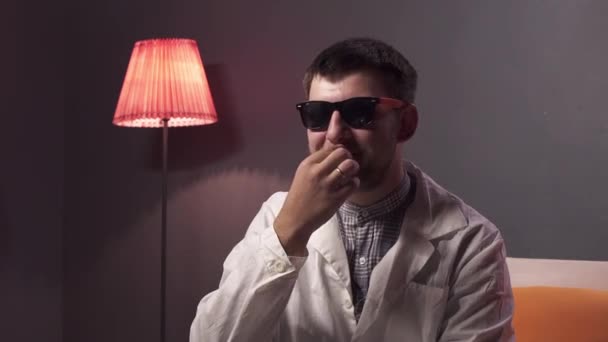 Mladý roztomilý muž s vousy nosí sluneční brýle a bílý laboratorní plášť toches knírek — Stock video