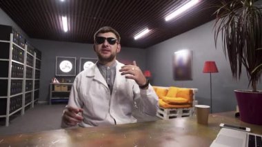 Güneş gözlüğü ve beyaz laboratuvar önlüğü giyen adam masaya kumbara yerleştirerek konuşuyor