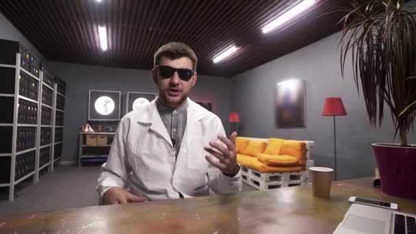 Young male in sunglasses and white lab coat speaks placing piggy bank on desk — Stockvideo