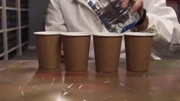 Hombre con bata de laboratorio blanca vierte agua de la botella de plástico a cuatro tazas de papel — Vídeo de stock