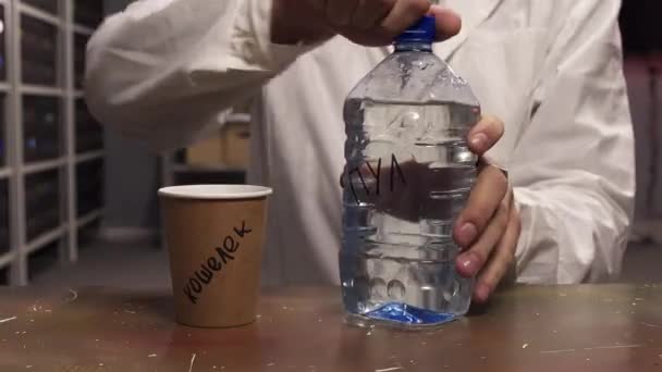 Muž oblečený v bílém laboratorním plášti nalévá vodu z plastové láhve na hnědý papírový pohár — Stock video
