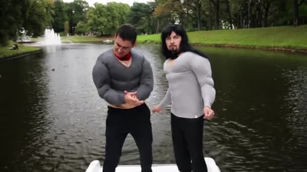 Två attraktiva män i falska muskler bröst och armar vadderade kostymer stå i båten — Stockvideo