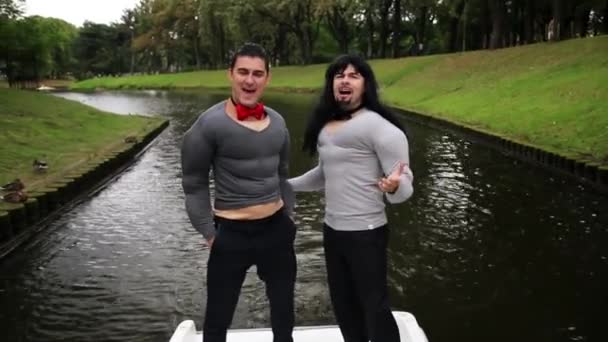 Két csinos fiúk viselt hamis izmok párnázott jelmezek tánc és énekelni a csónakban — Stock videók