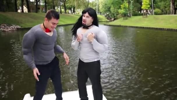 Zwei kaukasische Männer in falschen muskelgepolsterten Hemden tanzen und singen im schwimmenden Boot — Stockvideo