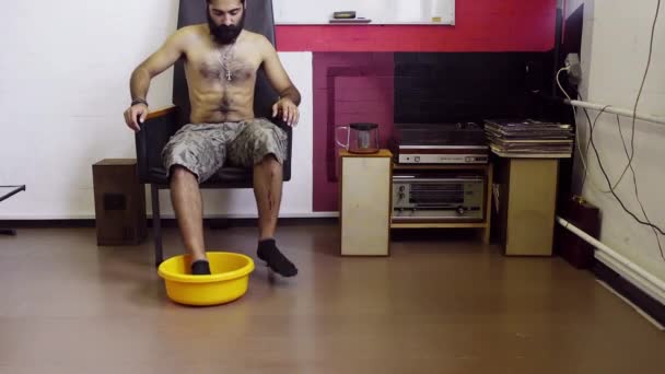 Hindu aussehender Kerl mit nacktem Oberkörper sitzt auf Stuhl, stellt Füße in gelbes Waschbrett — Stockvideo