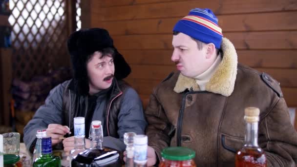 Twee mannen dragen winterkleren en hoeden zitten rond tafel met eten en praten — Stockvideo
